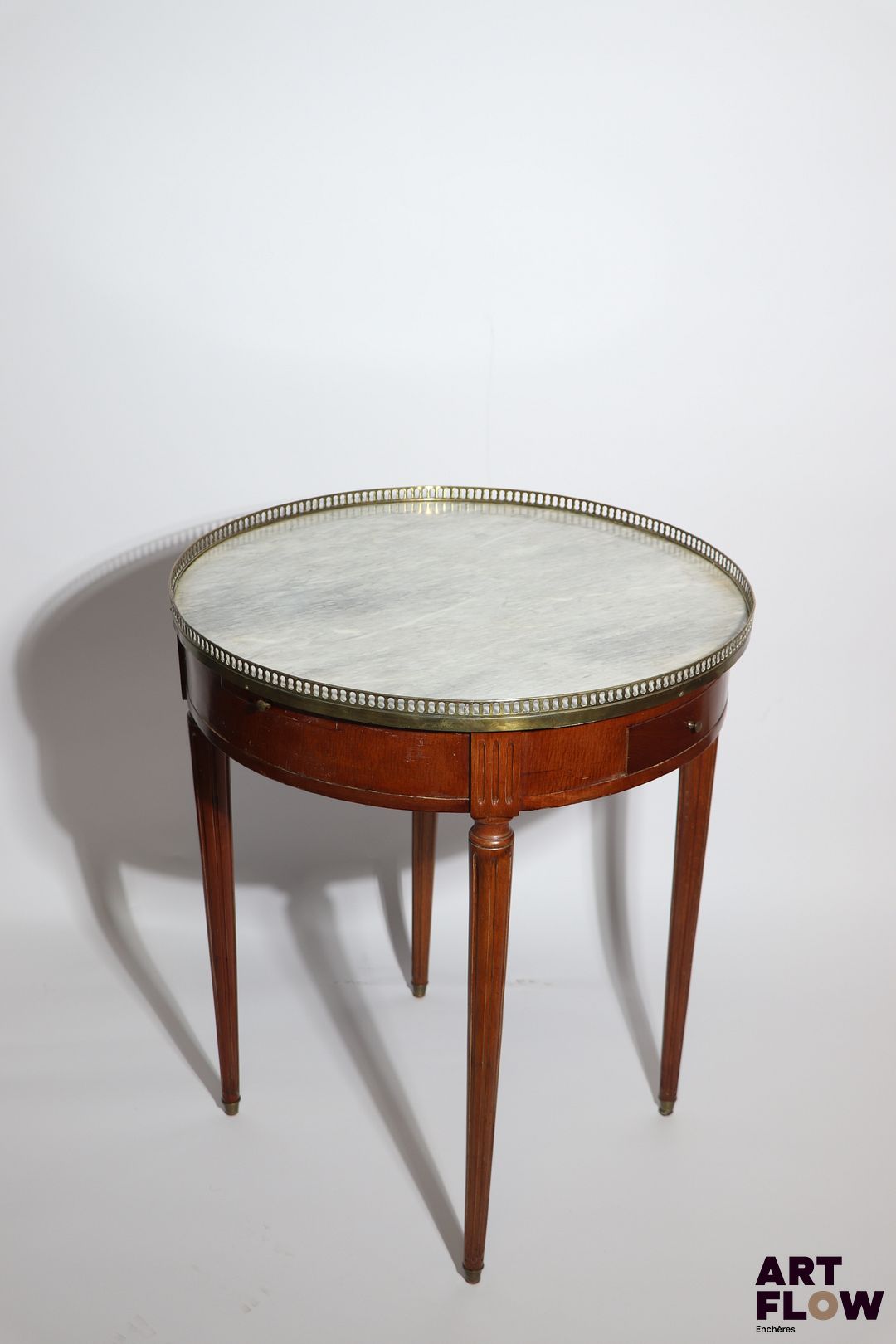 Table bouillotte en bois de placage, posant sur pieds fuselés et cannelés, le plateau en marbre blanc entouré d'une galerie de laiton ajouré. Style Louis XVI. Petits accidents. H 74, d 65 cm Facture de réparation de mai 2011.