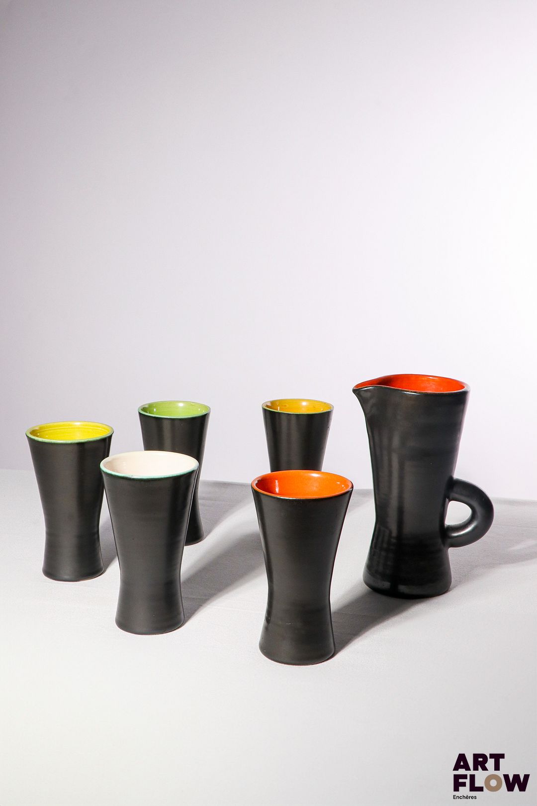 POL CHAMBOST (1906-1983) Service à orangeade 860, comprenant un pichet et 5 verres de forme diabolo, en faience émaillée à couverte noire à l'extérieure et colorée à l'intérieur (jaune, vert et orange). Le pichet et les verres tamponnés en creux "860, Made in France" H. pichet : 19,5 cm H. verres ; 14 cm Bibliographie : Philippe Chambost, "Pol Chambost, Sculpteur-Céramiste 1906-1983", Somogy Editions d'Art, Paris, 2006, modèle similaire p. 115 ; p. 145