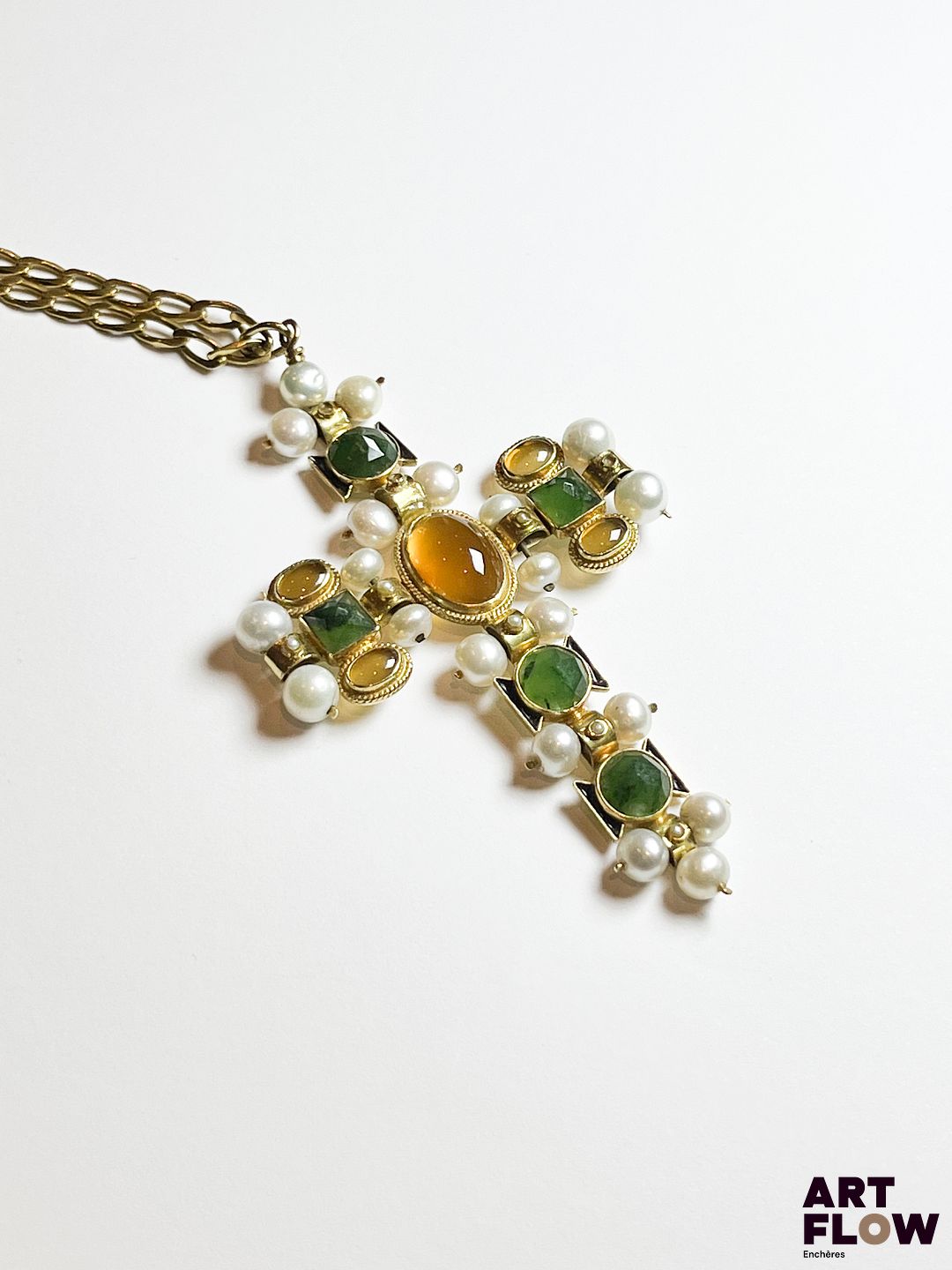 Croix en or jaune 750‰, portant des poinçons italiens et français, de style étrusque, composée au centre d’une belle citrine cabochon sur une frise enrubannée, la croix représentée par un ensemble de perles blanches, petites citrines cabochons et probablement des néphrites de forme carrée facettées, soulignées par de l’émail noir. La croix suspendue sur une chaine en or jaune 750‰ massive, maille cheval avec chainette de sécurité. Poids : 32.29 g brut Dimensions croix : 8 x 4.5 cm – Longueur chaine : 59 cm Expertise : Philippine Vanheems