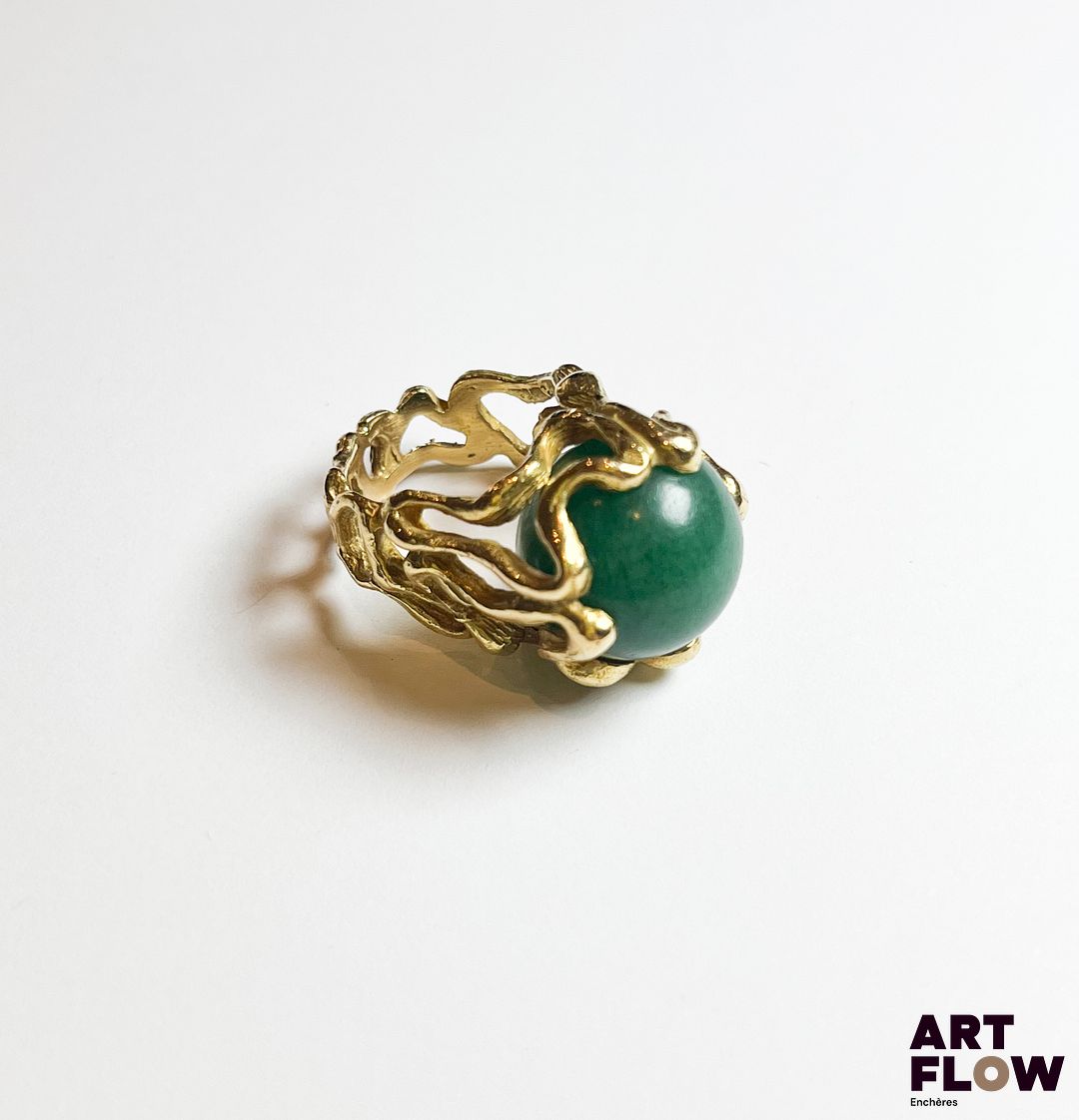 Bague en or jaune 750‰, poinçon tête d’aigle, composée d’un corps entièrement contourné et ajouré, au centre repose une aventurine verte de forme sphérique dans le style de Gilbert Albert. Légères fissures sur le corps Poids : 15.12 g brut Tour de doigt : 48 Expertise : Philippine Vanheems