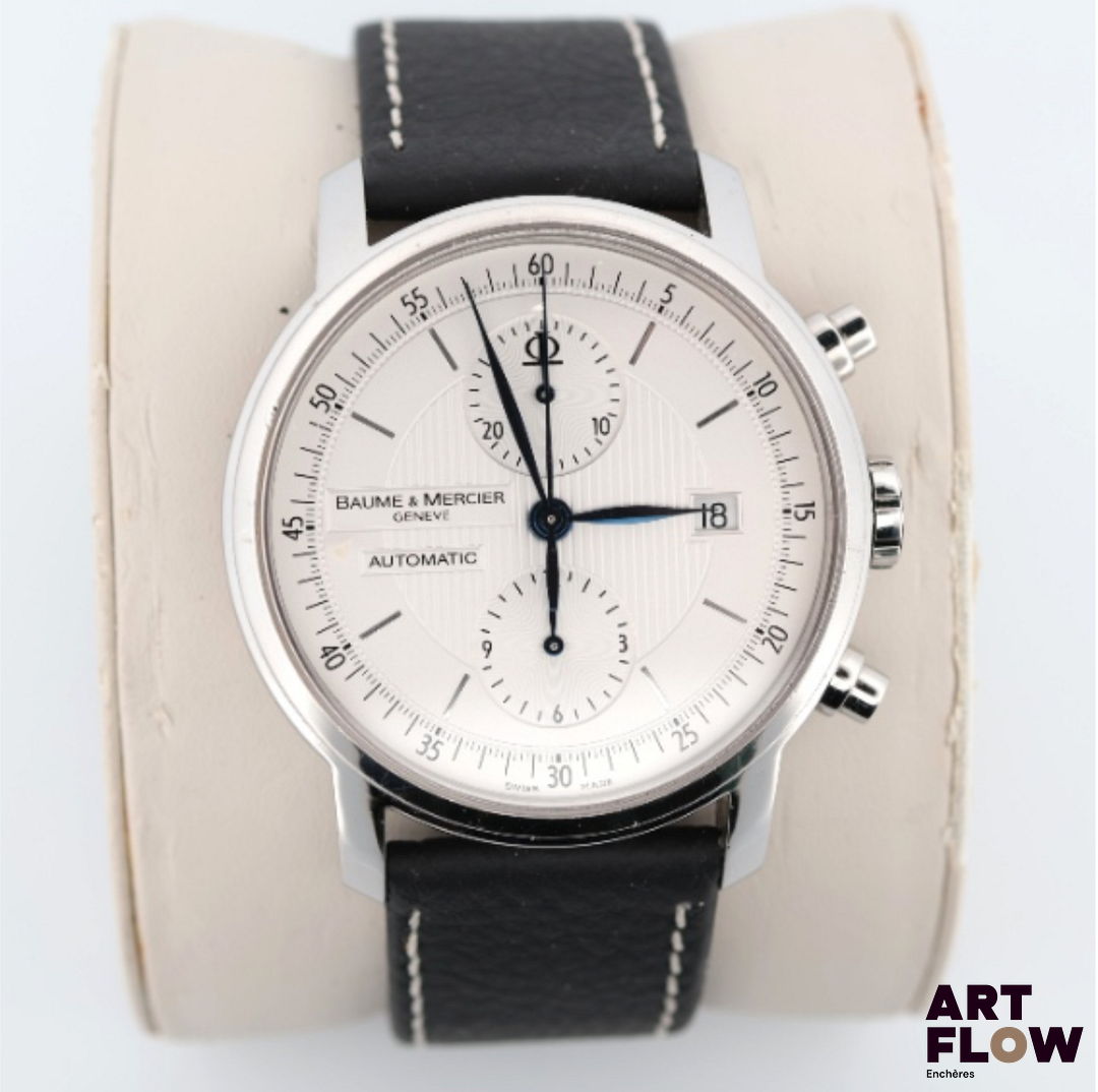 BAUME & MERCIER Classima, Référence 65560 - Montre bracelet chronographe en acier, boitier rond, ouverture au doslaissant apparaître le mouvement, signé. Cadran argenté, index bâtons et aiguilles feuilles, 2 compteurs pour la fonction chronographe. Mouvement automatique cal. BM 1377750, signé. Bracelet en cuir neuf mais boucle d’origine signée dans l’écrin. Diamètre 42mm Accompagné d’une carte de garantie du 03/10/2012 tamponnée « Elysées Horlogerie » à Paris, écrin et sur-boîte, manuel d’utilisation Mouvement opérationnel au moment de la mise en catalogue, mais révision conseillée. Bon état général, quelques rayures d’usage, cadran préservé.