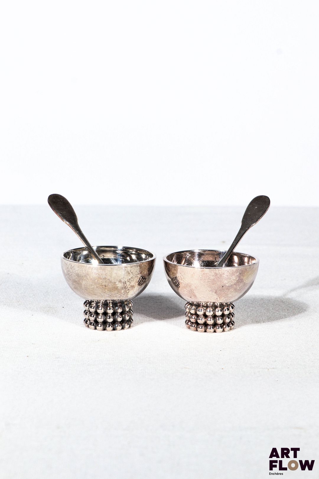 Jean DESPRES (1889-1980) Paire de salerons en métal argenté sur talon orné d'un triple rang de perles. Avec deux petites cuillères. Signé "J.Despres' à la pointe sur le corps. H. 3 x Diam. 4 cm Petites rayures d'usage.