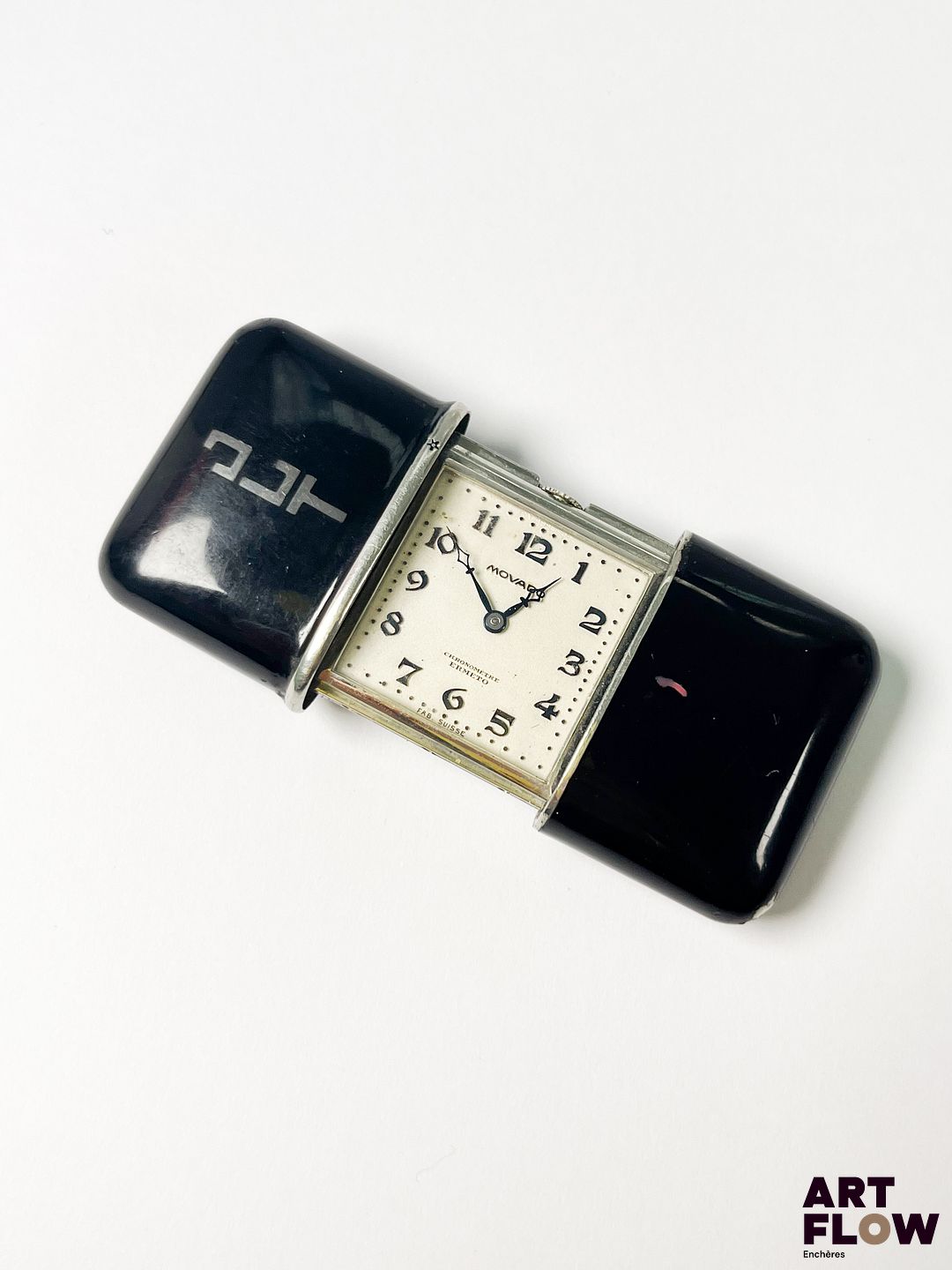 MOVADO Modèle Ermeto Montre de voyage en bakelite, à mouvement mécanique à remontage glissière, le boîtier rectangulaire à cadran à fond argenté, aiguilles losange bleuies, index appliqués en chiffres arabes bleuies Années cira 1930 Dimensions : 33 x 43 mm Bon état général Monogrammée « JLT » Fonctionnelle au moment de la rédaction du catalogue, mécanisme non garanti. La Movado Ermeto est lancée dans les années 1920 par la marque suisse Movado, conçue comme une montre de voyage compacte et élégante. Elle se distingue par son boîtier coulissant protecteur et son style Art déco, devenant un accessoire prisé pour sa robustesse et son raffinement. Expert : Monsieur Alexis Francis-Boeuf