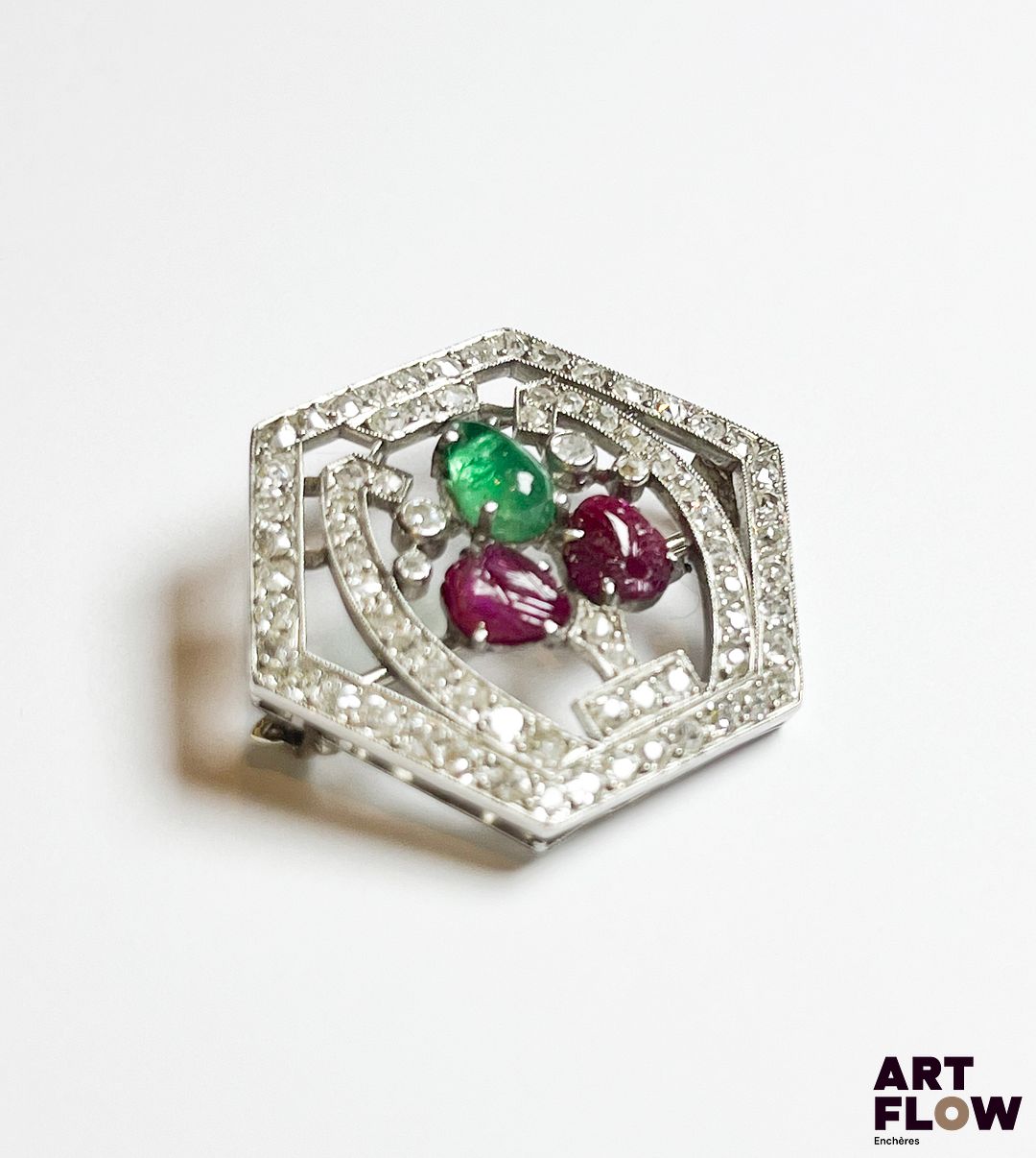 Broche en platine et épingle en or blanc 750‰ (poinçons tête d’aigle et tête de chien), de forme hexagonale, en forme de bouquet de fleurs dans un cadre géométrique, délicatement ajouré, entièrement pavé de diamants taille ancienne ; le bouquet représenté par une émeraude poire cabochon et deux rubis sculptés dont un percé. Bouquet dessoudé sur deux points à l’arrière. Poids : 7.16 g brut Dimensions : 2.4 x 2.9 cm Expertise : Philippine Vanheems
