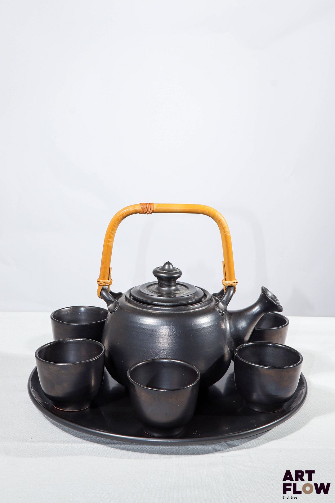 JEAN MARAIS Service à thé en céramique à glaçure noire comprenant : - 1 plateau rond. D. 32 cm - 1 théière avec anse en osier (petite egrenure au bec verseur). Signée. H. 21,5 cm - 6 tasses. H. 6 cm Très bon état général.