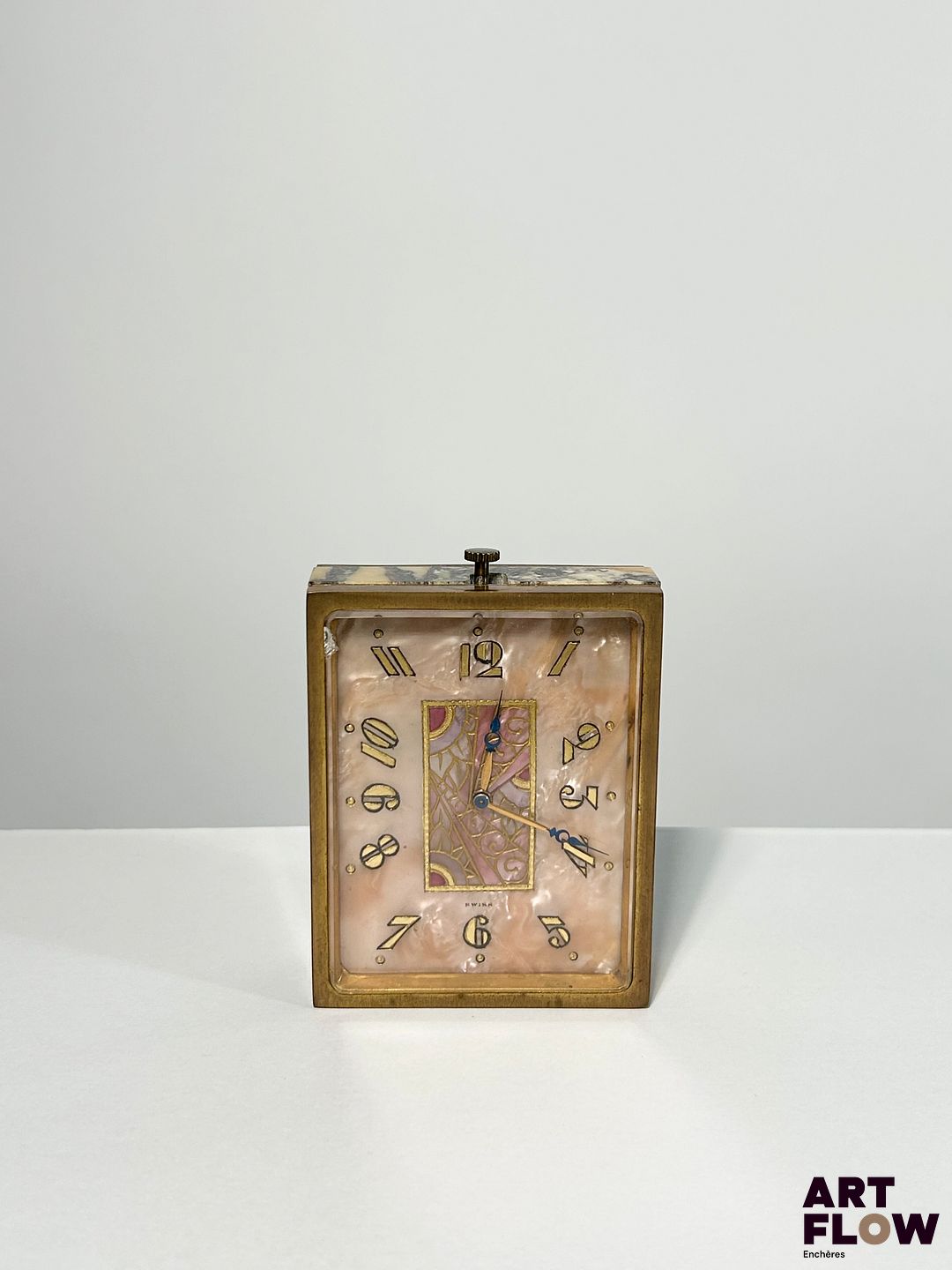 JUVENIA Pendule flacon en bronze doré et marbre jaune, cadran en marqueterie de nacre, aiguilles Breguet, index en chiffres arabe peints, signée au dos 8 x 6 cm Bon état général (absence du bouchon initialement sur le remontoir) Juvenia est une manufacture horlogère suisse fondée en 1860 à La Chaux-de-Fonds. Elle s’est illustrée par des montres élégantes et innovantes, notamment des boîtiers à ouverture frontale et des mouvements de haute qualité. Au XXᵉ siècle, Juvenia devient une marque reconnue internationalement, symbole du raffinement horloger suisse.