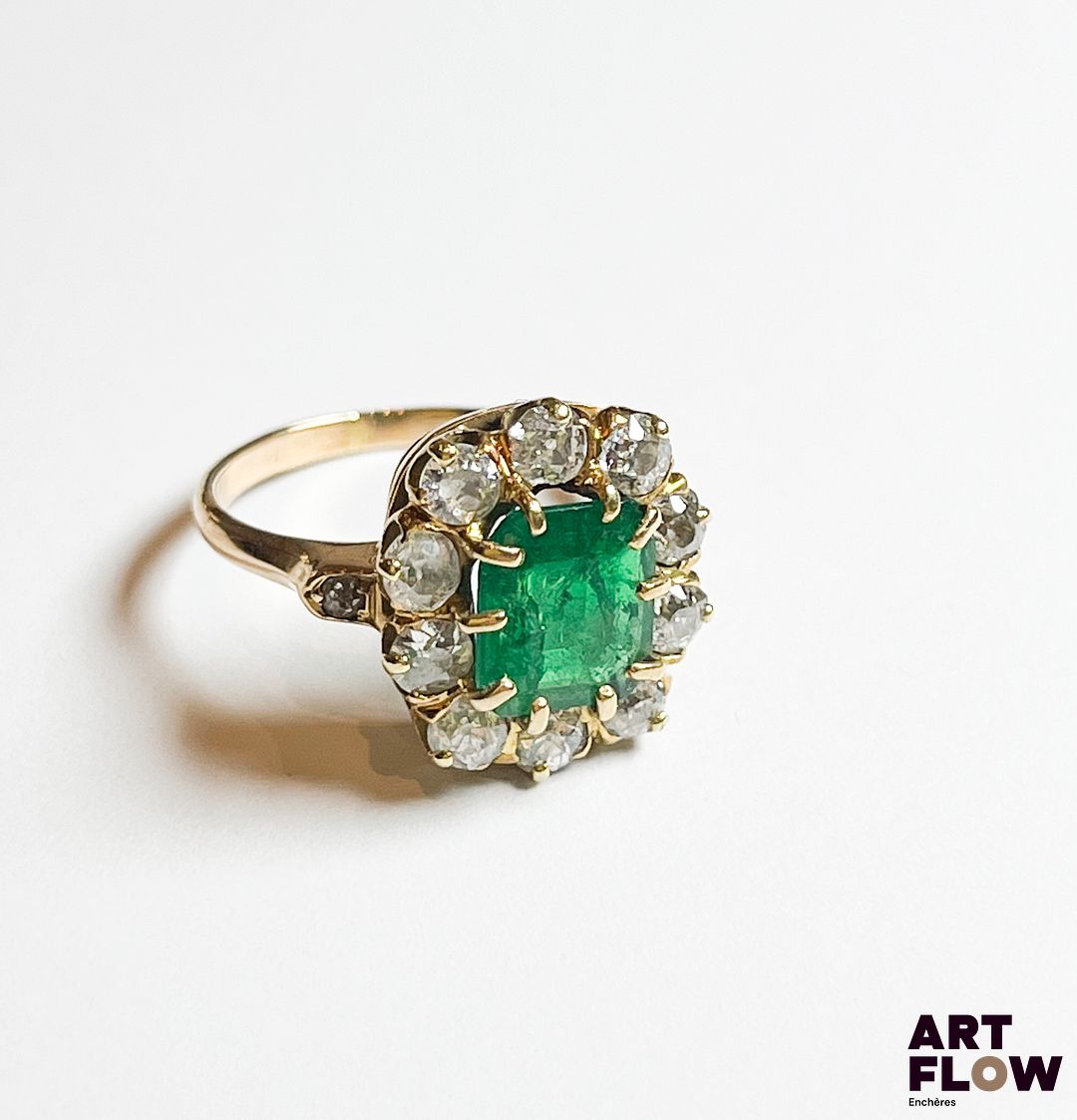 Bague en or jaune 750‰, poinçon hibou, de forme marguerite, sertie au centre d’une émeraude rectangulaire à pans coupés d’un magnifique vert (8.3x7.5mm), et entourée de 10 diamants taille ancienne ; le panier délicatement ajouré, le corps également mis en valeur par deux petits diamants. Quelques manques et accidents à l’émeraude. Poids : 4.62 g brut Diamants : 1 carat env. – Emeraude : 1.2 carat env. Tour de doigt : 52 Expertise : Philippine Vanheems